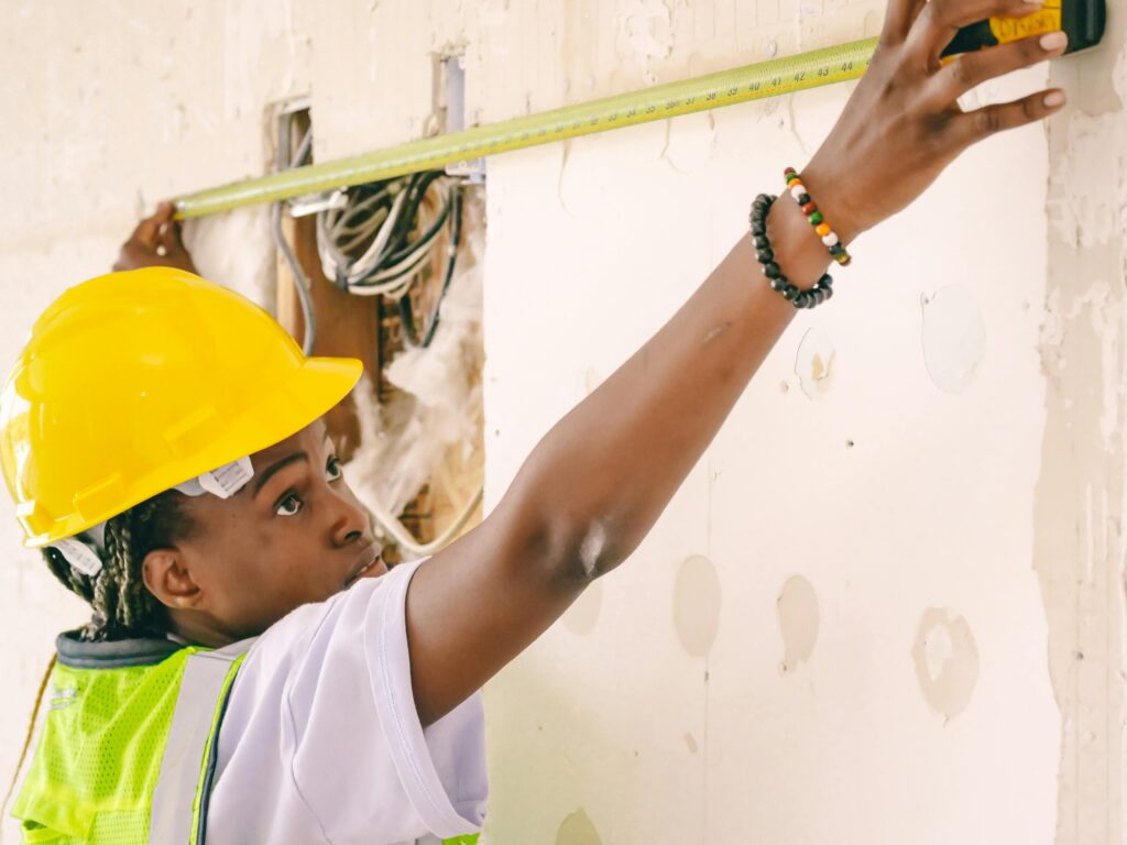 Black woman contractor wearing a yellow hard hat and safety vest, measuring a wall with precision, demonstrating cost control strategies for contractors.