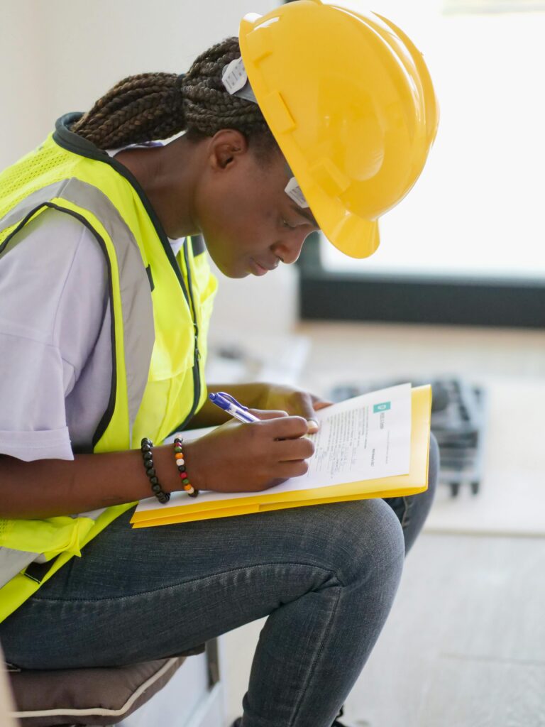  Black woman contractor in a yellow hard hat and safety vest, reviewing documents on a construction site, focusing on managing construction expenses effectively.